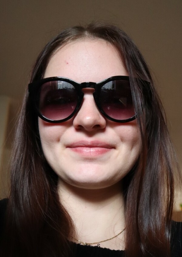Person mit langen braunen Haaren und einer großen runden Sonnenbrille, lächelnd im Innenraum.